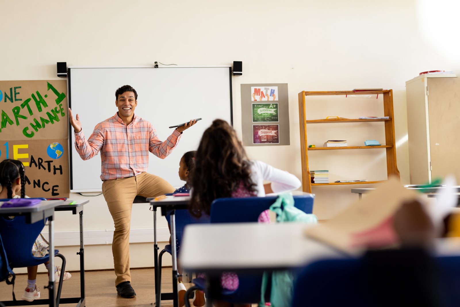Diverse happy male teacher with tablet and elementary schoolchildren sitting at desks in class. Technology, education, childhood, development, learning and elementary school, unaltered.