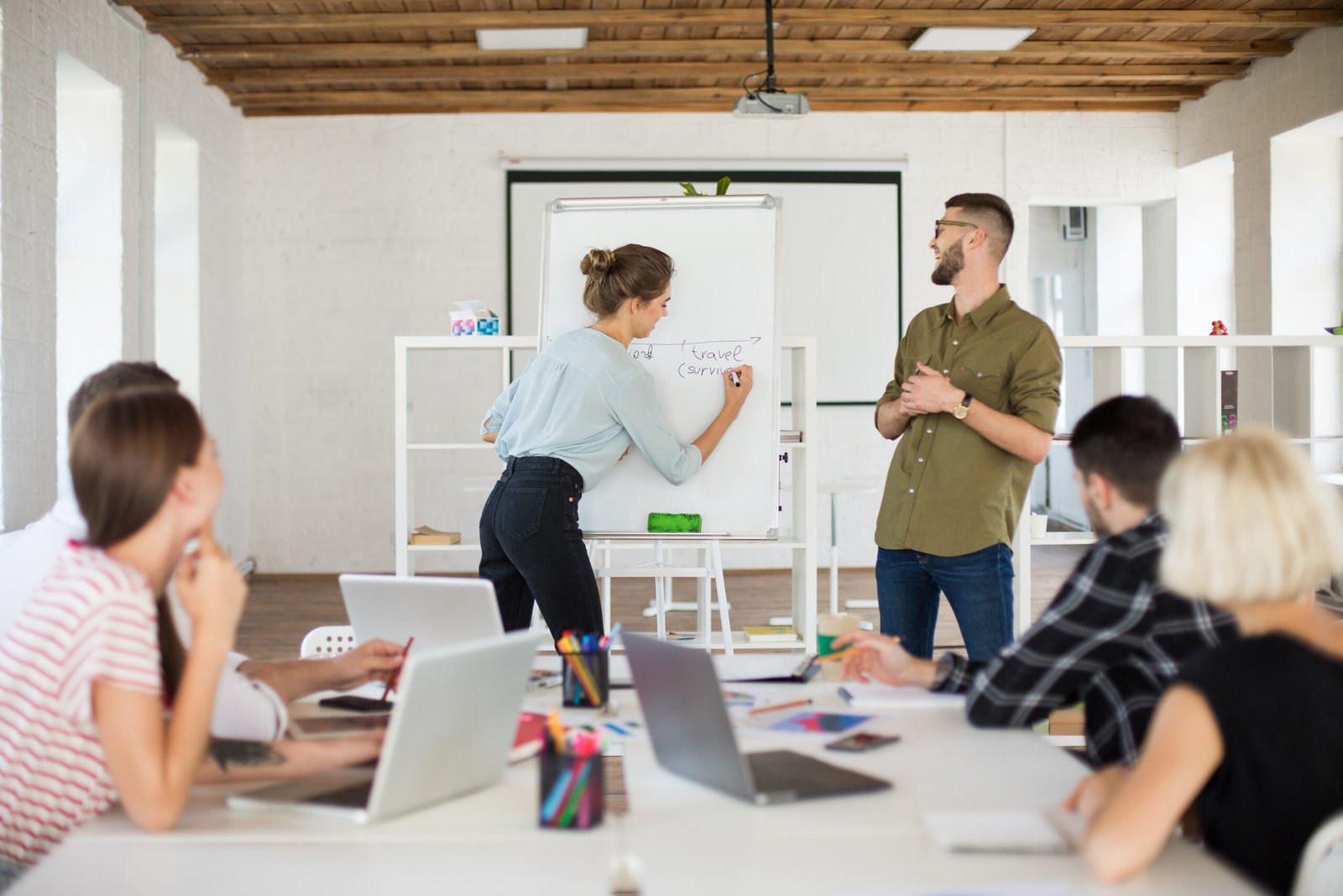 Young man in eyeglasses and shirt and pretty woman in blouse standing near board happily presenting new project to colleagues. Group of creative people working together in modern white office
